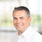 Ein Mann mit kurzen dunklen Haaren und weißem Kragenhemd lächelt in die Kamera. Der Hintergrund ist mit natürlichem Licht und einem Hauch von Grün sanft verschwommen.