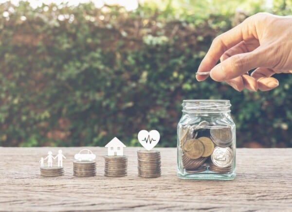 Eine Hand lässt eine Münze in ein mit Münzen gefülltes Glasgefäß fallen. Davor befinden sich vier Münzstapel mit Symbolen: eine Familie, ein Auto, ein Haus und ein Herz, die das Sparen für verschiedene Lebensziele symbolisieren.