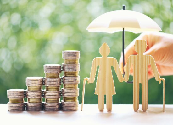 Münzstapel neben den Holzfiguren eines älteren Ehepaars mit Blindenstöcken, während eine Hand einen Regenschirm über sie hält, als Symbol für finanziellen Schutz und Sicherheit im Ruhestand.