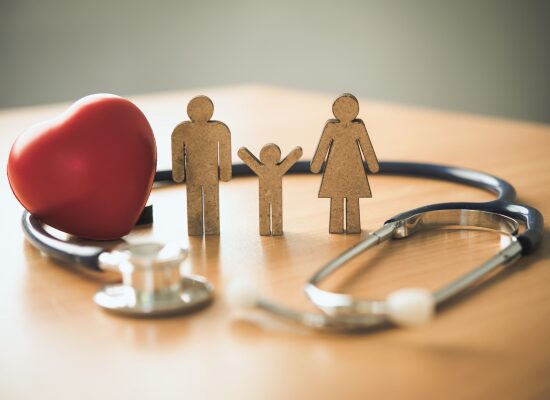 Holzfiguren einer Familie mit einem Kind stehen neben einem roten Herz, umgeben von einem Stethoskop auf einem Holztisch, das Gesundheit und Familienpflege symbolisiert.