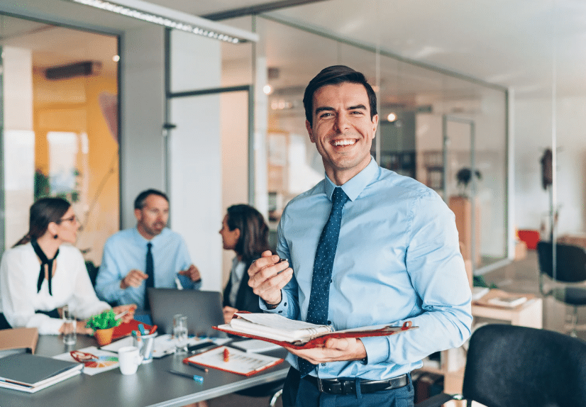Ein lächelnder Geschäftsführer in blauem Hemd und Krawatte hält ein Klemmbrett in einem modernen, hellen Büro mit Glaswänden, während drei Kollegen an einem Tisch im Hintergrund über Pensionszusagen diskutieren.